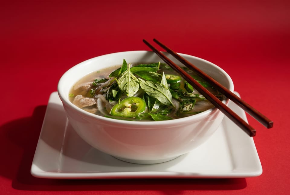 A bowl of pho with basil, jalapenos, and chopsticks on a white plate against a red background.