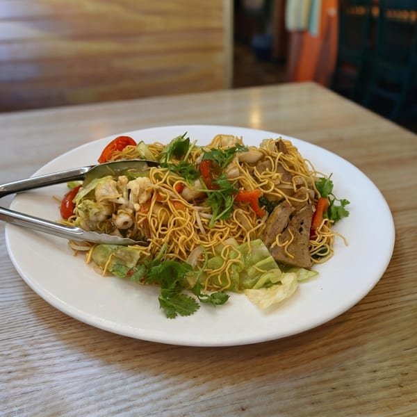 A plate of stir-fried noodles with vegetables, meat, and seafood, with serving tongs.