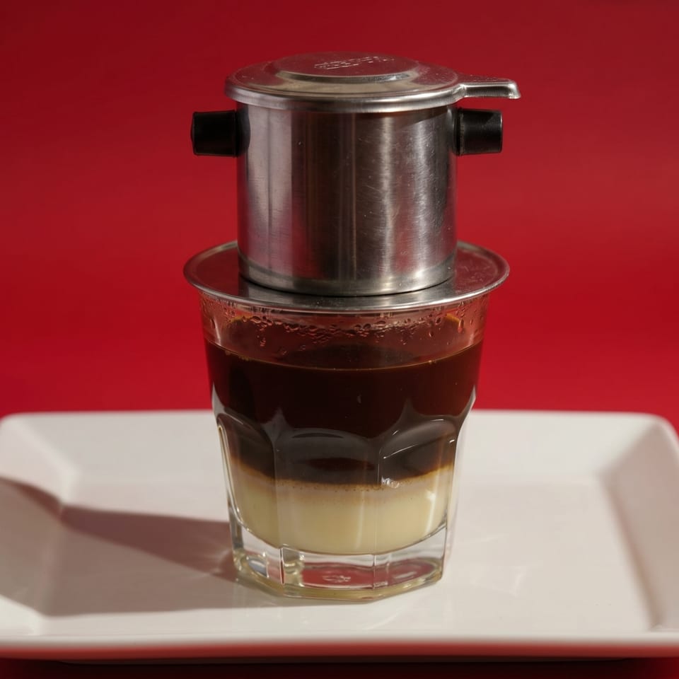 Vietnamese coffee in a glass with a metal phin filter on a white plate against a red background.
