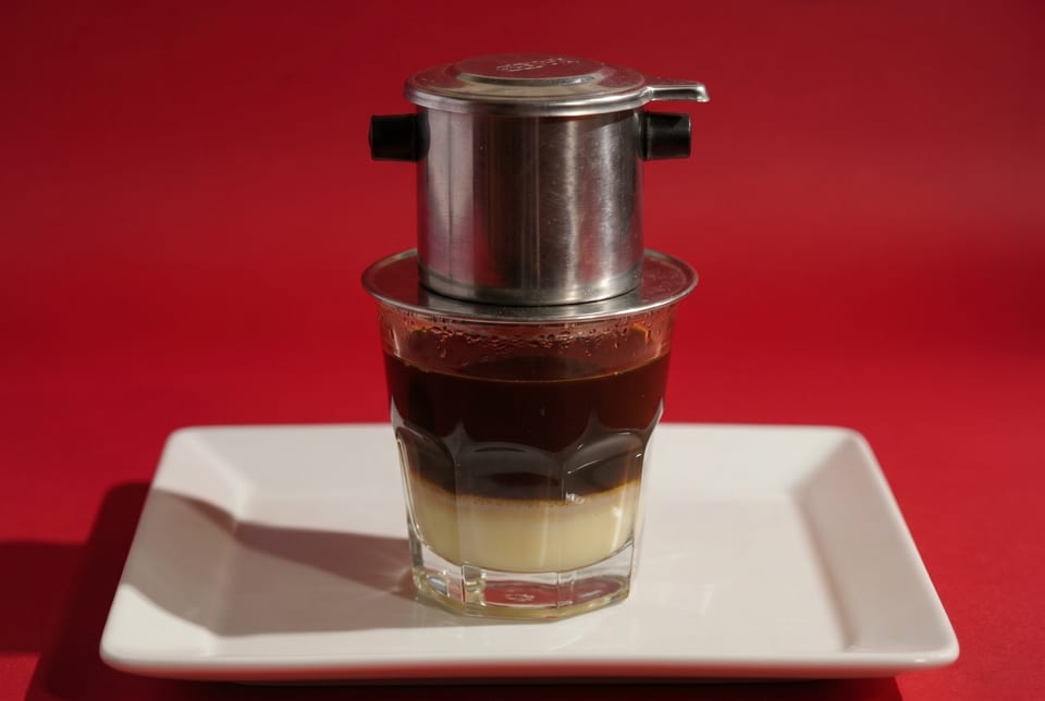 Vietnamese coffee in a glass with a metal phin filter on a white plate against a red background.