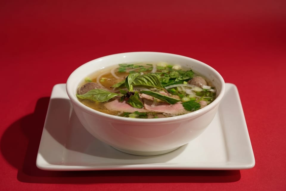 A bowl of Vietnamese pho with beef, noodles, and herbs on a white plate against a red background.