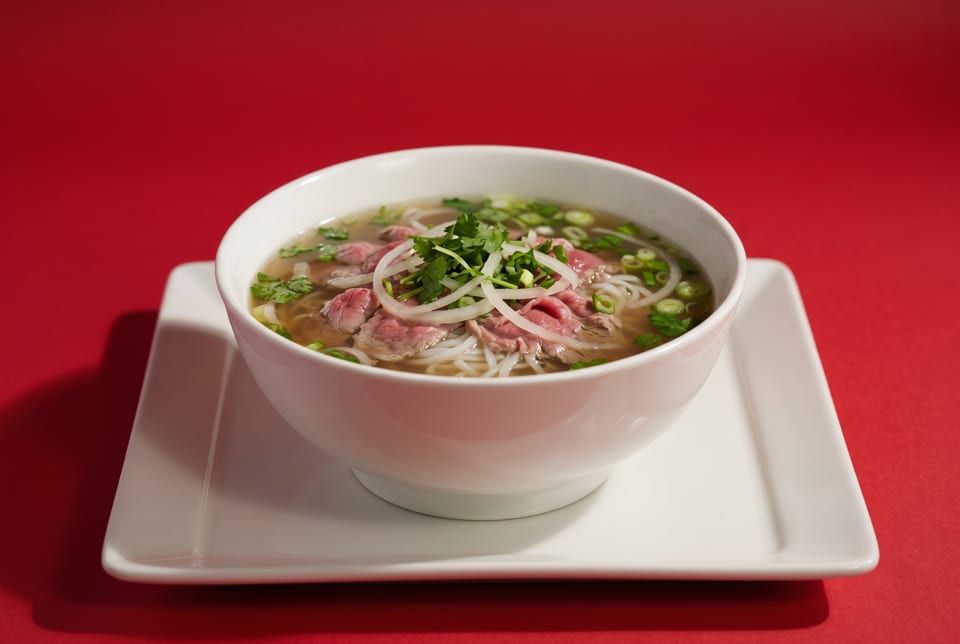 A white bowl of pho with beef, noodles, onions, and herbs on a white plate against a red background.